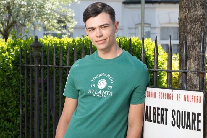 Will Mitchell, in a green t-shirt, stood in front of the Albert Square sign in EastEnders