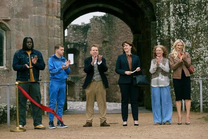 A group of people stand in a row at a ribbon cutting ceremony at the entrance of a historic castle. Some are smiling, while others appear disinterested.