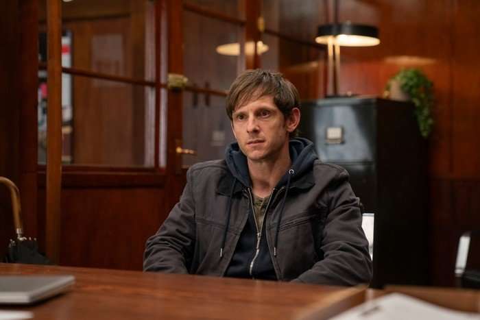 Jamie Bell as Niall in Half Man, sat at a wooden desk in an office, wearing a grey jacket over a blue hoodie.