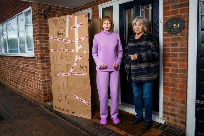 Linda stands next to her 'Ann Droid' cardboard box, outside the front door of Sue's home. Sue's stands next to her, looking concerned.