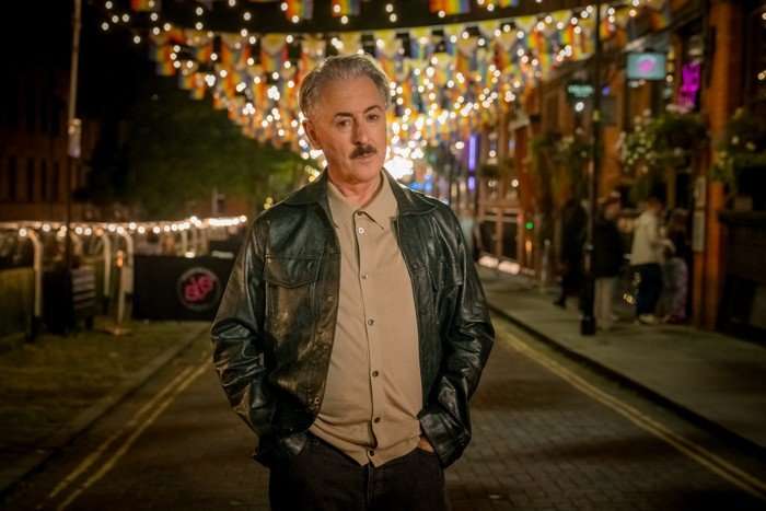 Leo (Alan Cumming) Leo (Alan Cumming) in Tip Toe; the character stands in a leather jacket and shirt on a street with pride flags and fairy lights hanging from the buildings