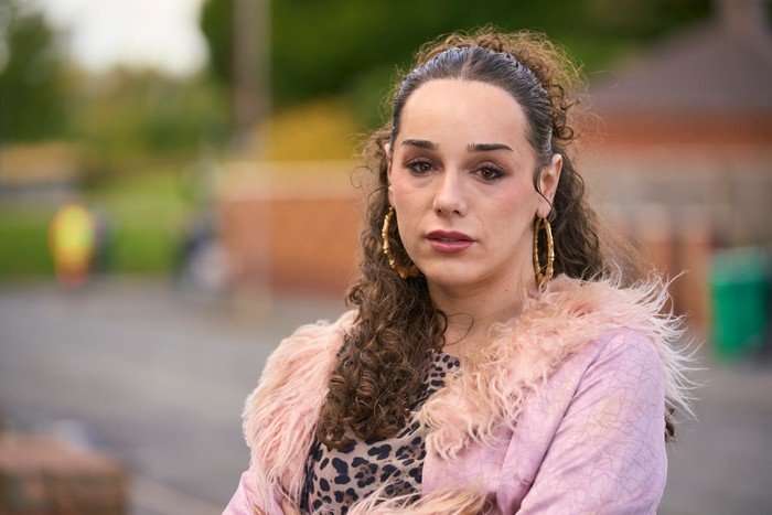 Iz Hesketh as Zee in Tip Toe. She is wearing a pink outfit and standing on a residential street in the daytime, looking concerned