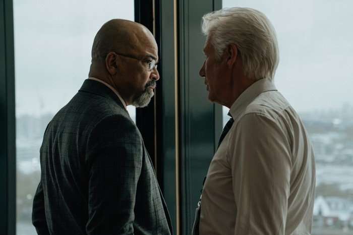 Two men stand by a window in an office building, having a serious discussion. Both are in suits, but Bosko (on the right) has his jacket off.