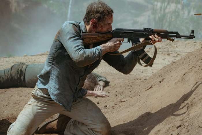 Michael Fassbender is in a ripped button up shirt and cream trousers, aims a machine gun at an unknown target in the desert
