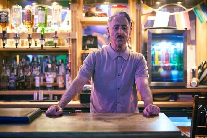 Alan Cumming as Leo Struthers in Tip Toe, standing behind a bar with bottles on shelves and in a fridge in the background.
