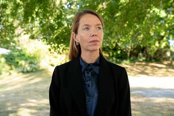Anna Maxwell Martin as Leanne in A Good Girl's Guide to Murder Anna Maxwell Martin as Leanne in A Good Girl's Guide to Murder, wearing a dark suit and standing outside under green trees on a sunny day.