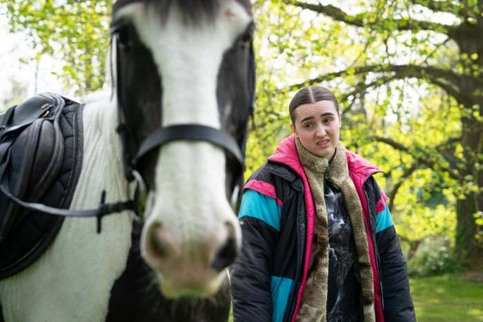 A young woman stands outdoors beside a black-and-white horse, which is close to the camera and slightly out of focus in the foreground. She wears a colourful padded jacket and a scarf, with a mildly unimpressed expression, while trees and greenery fill the background.