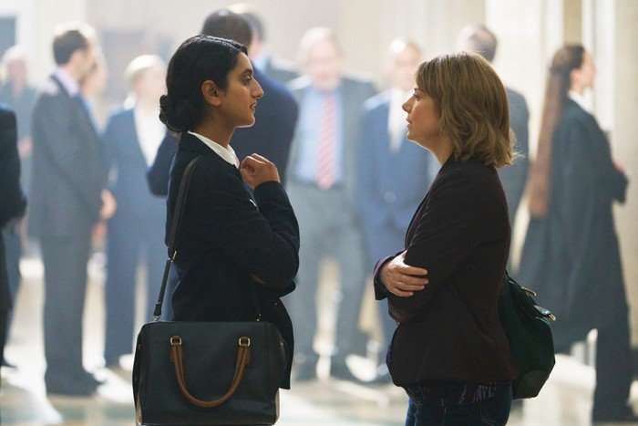 Two women stand facing one another in a busy courthouse corridor, deep in conversation as blurred figures move around them in the background. Dressed in dark formal clothing and carrying bags over their shoulders, the pair appear tense and focused amid the subdued, high-pressure atmosphere.