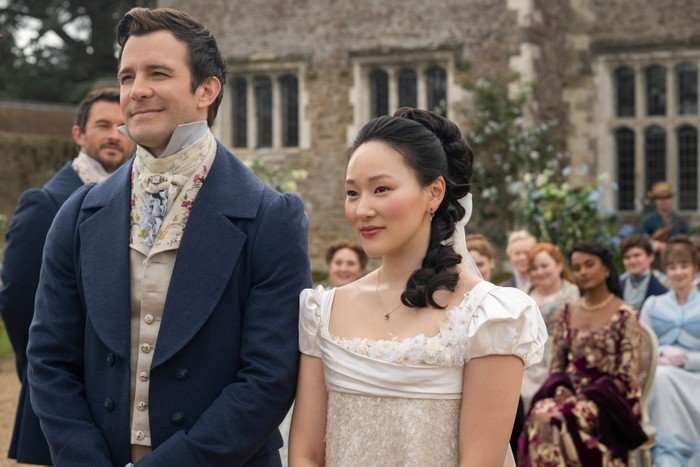 Bridgerton-s4-p2-YerinHa Luke Thompson as Benedict Bridgerton, Yerin Ha as Sophie Baek in Bridgerton standing at the end of the aisle in wedding attire before the priest, smiling.
