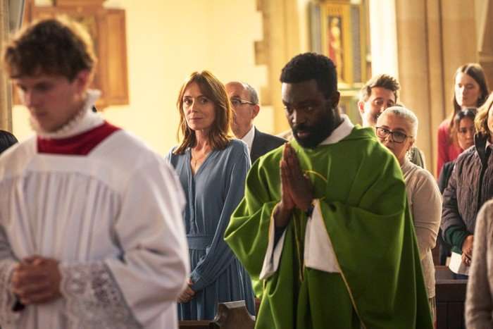 A church congregation stands during a service as a priest in green vestments walks down the aisle with hands clasped in prayer. In the background, a woman in a light blue dress watches solemnly, surrounded by other attendees inside the softly lit church.