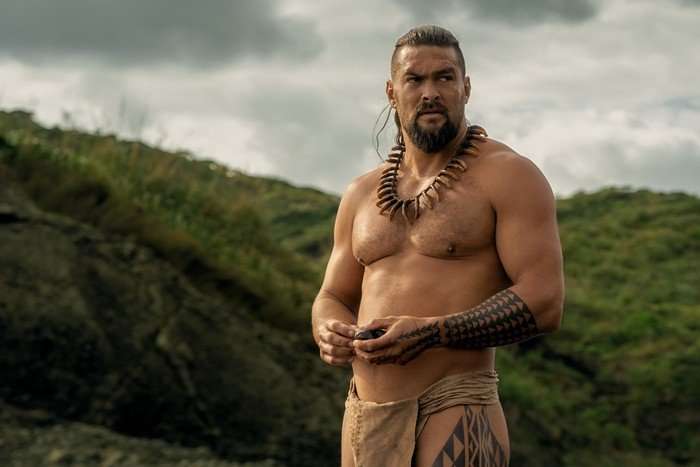 Chief_of_War_Photo_010104 Jason Momoa stars in Chief of War; he is seen here shirtless against a cloudy backdrop with large green hills, looking stern and deep in thought
