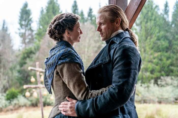 A couple in period clothing stand close together under a wooden shelter in the rain, looking into each other&rsquo;s eyes with a forest behind them.