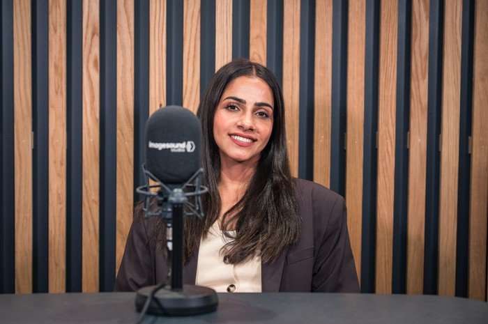 Mandip Gill sat behind a desk with a microphone, looking at the camera and smiling