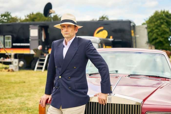 A man wearing a cream fedora and a navy double-breasted blazer leans against the front of a red Rolls-Royce parked in a grassy field. He looks serious as trailers and equipment can be seen in the background, suggesting a filming location or event setting. Trees and a cloudy sky are visible behind him.