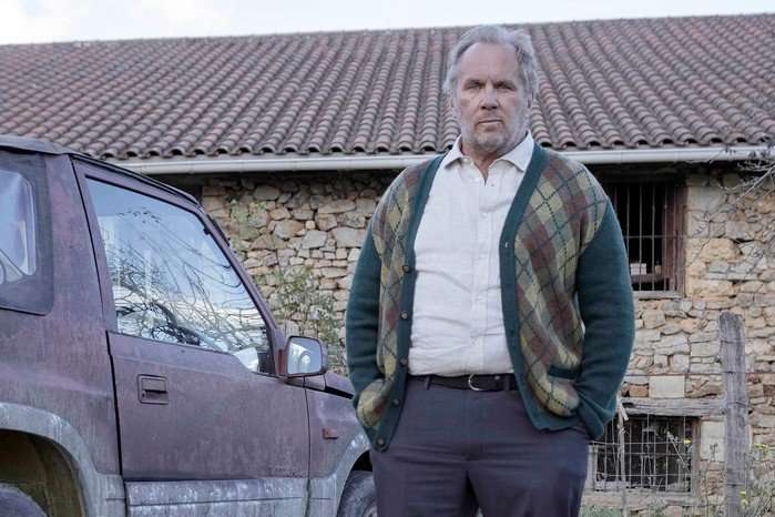 A man stands outdoors beside an older, slightly dirty SUV parked in front of a rustic stone building with a tiled roof. He appears middle-aged, with short greying hair and light stubble. He&rsquo;s dressed in a white button-up shirt and a patterned cardigan with a diamond (argyle-style) design, and his hands are tucked into his pockets. The setting looks rural, with a simple fence and some sparse vegetation nearby. His expression is serious and contemplative, and he faces forward as if watching or waiting for something.