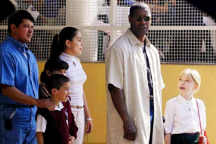 A man wearing sunglasses stands beside a young blonde girl in a school uniform, looking down at her as she looks up at him. Nearby, a family with two children stands together, watching the interaction. They appear to be outside a school, with a yellow wall and metal fencing in the background.