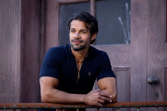 Junade Khan as Idris Nazir in Coronation Street, stood in front of a burgundy door and leaning on a rusty metal fence. He is wearing a blue top which is zipped open.