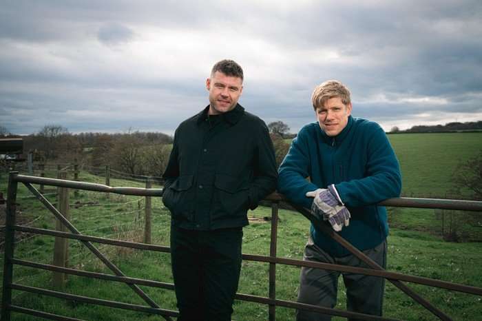 EMBARGO_EMMERDALE_01 Aaron and Robert stood against a fence on open farmland in a scene from Emmerdale