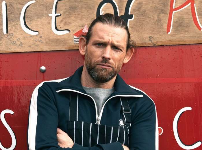 Chris Coghill as Kev Townsend poses against a red wall in an apron with the sign behind saying "A Slice of Kev-en...Cakes and Coffee' in Emmerdale.