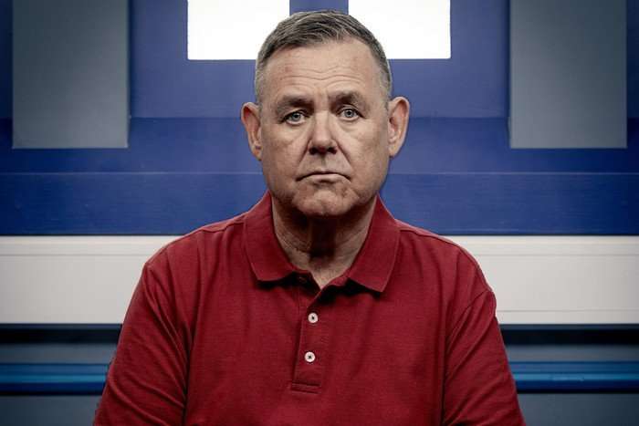 George Shuttleworth, wearing a red polo shirt, sits in a police interview room in Coronation Street