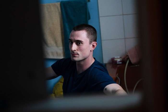 A Good Girl's Guide to Murder series 2 A candid portrait of a young man with a very short haircut sitting indoors in what appears to be a bathroom or small tiled room. He is wearing a dark navy T-shirt and looking toward the camera with a serious, alert expression. The scene is framed through a narrow gap or doorway, creating a voyeuristic, intimate perspective. Blue walls, hanging towels, and tiled surfaces are visible in the softly lit background, giving the image a quiet, tense atmosphere.