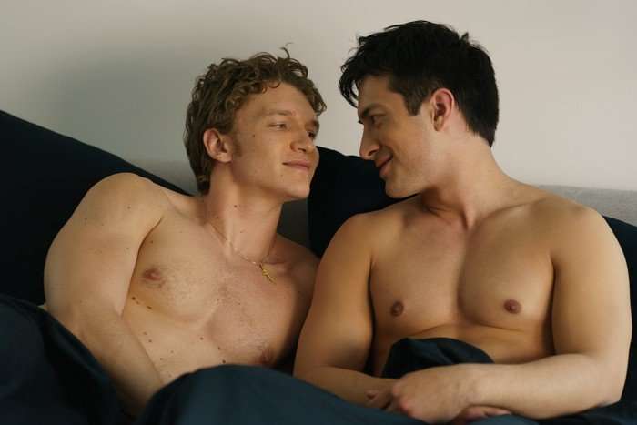 Heated Rivalry Ilya Rozanov (Connor Storrie) and Shane Hollander (Hudson Williams) in Heated Rivalry; they are sat together shirtless in bed, and smiling as they look in each other's eyes
