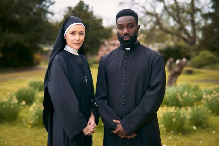 Falling A nun and a priest stand side by side outdoors in a garden or park setting. Both are wearing traditional black religious clothing. The nun wears a black habit with a white head covering and a cross necklace, while the priest wears a black cassock with buttons down the front and a clerical collar. They are facing the camera with calm, serious expressions. The background is softly blurred, showing trees, grass, and flowering plants in natural daylight.