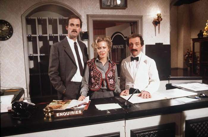 Fawlty Towers - Comedy Classic 06 John Cleese, Connie Booth and Andrew Sachs in Fawlty Towers. The trio are standing behind a reception desk of the hotel looking seriously at the camera. Cleese as Basil is in a dark grey suit, white shirt and tie and has a moustache; Connie as Polly is in a grey shirt and floral patterns vest, and Sachs as Manuel is in a white dinner jacket and black bow-tie, and also has a moustache.