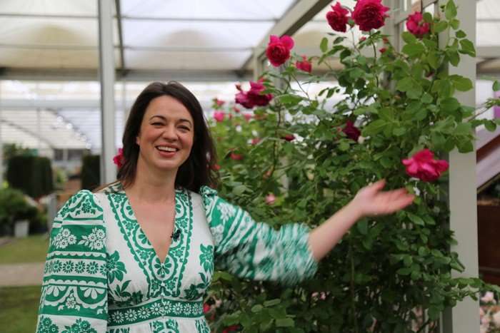 Frances Tophill wearing a green and white dress and standing in front of red flowers with green stems, smiling.