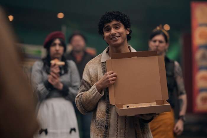 Anna Maryan as Justine, Bilal Hasna as Jesus and Andrew O'Neill as Mutt's Spouse in Good Omens. Jess is holding a pizza box and smiling while the others are stood behind him.
