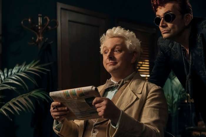 Michael Sheen as Aziraphale and David Tennant as Crowley in Good Omens. Aziraphale is sat down with a newspaper in his hands, while Crowley is leaning over his shoulder.