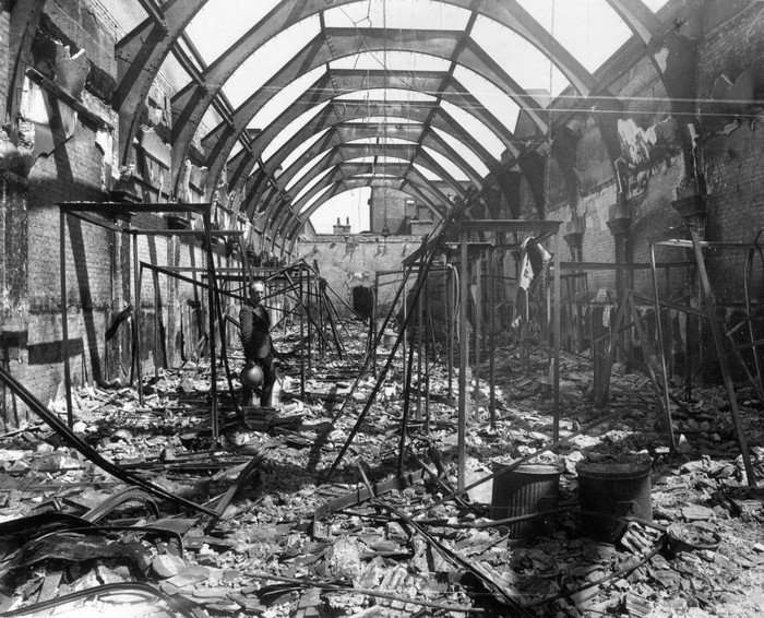 Black and white image of a man holding a helmet standing in the bombed out remains of a large building, now open to the sky. Twisted metal lies on the ground, while the steel roof structure remains.