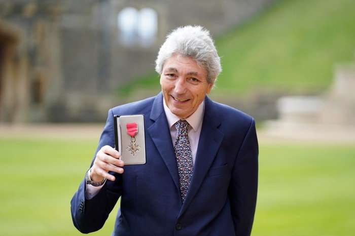 Paul Mayhew-Archer, wearing a suit and holding up his MBE.