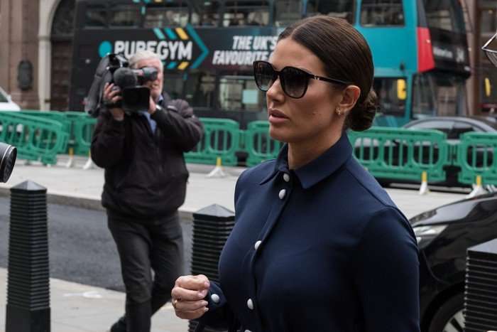 Rebekah Vardy in dark suit, hair tightly tied back and in sunglasses heads to court, with a photographer behind her.