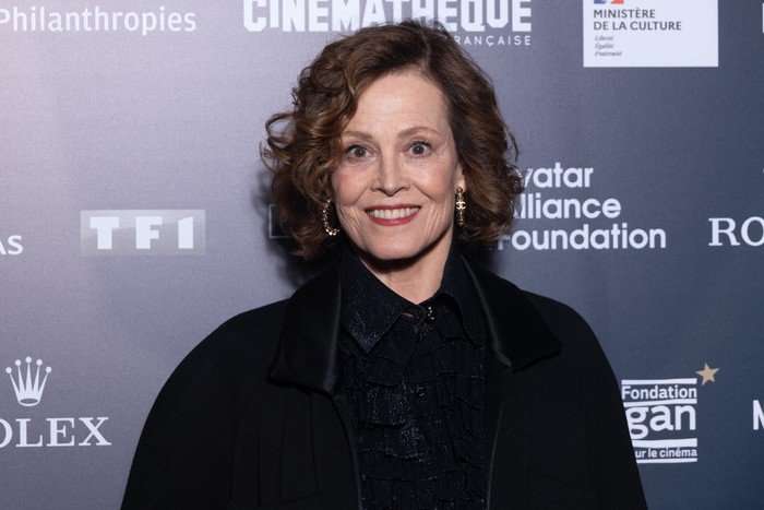 Sigourney Weaver smiling and standing in front of a blue backdrop