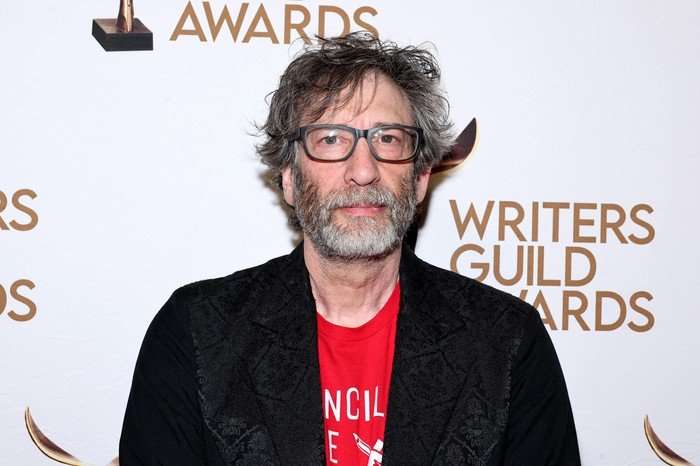 Neil Gaiman attends the 2024 Writers Guild Awards New York Ceremony in April 2024 wearing a red t-shirt and black blazer