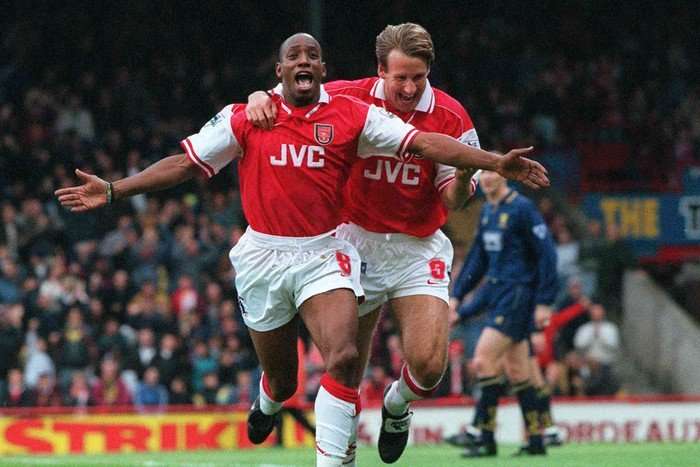 Arsenal striker Ian Wright, in red and white football, spreads his arms and smiles as he celebrates with Paul Merson.
