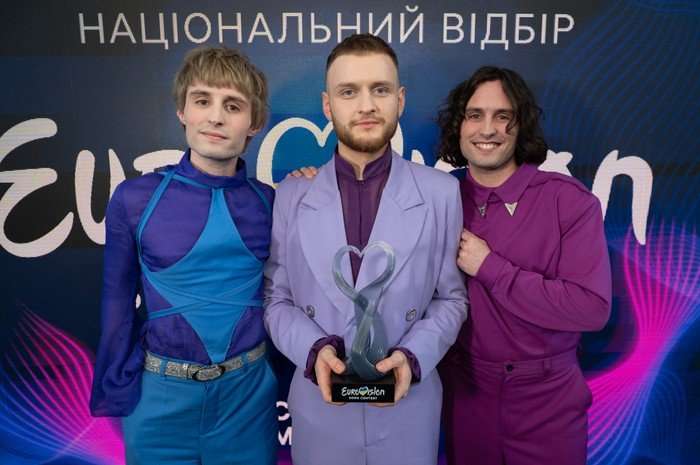 The three members of the Ziferblat stand shoulder to shoulder on the red carpet looking elated and smile at the camera. The band member stood in the middle holds in his hands a Eurovision trophy made of glass in the shape of a heart.