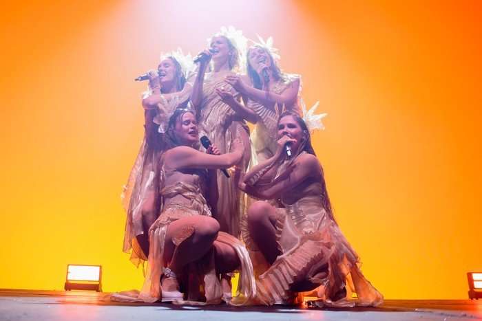 Tautumeitas performs on stage wearing matching gold dresses and head pieces. They cluster around each other whilst singing, three standing in the back and two crouching before them.
