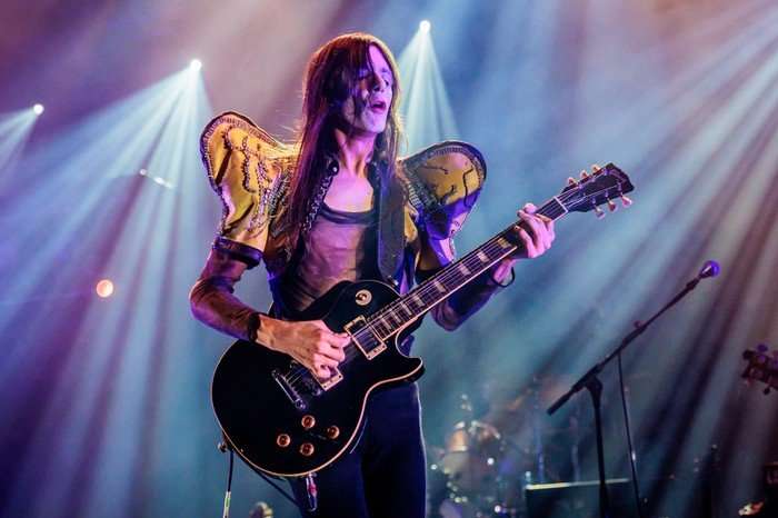 Lucio Corsi plays guitar on stage wearing a sheer top with over sized yellow shoulder pads with his long hair partially obscuring his face.