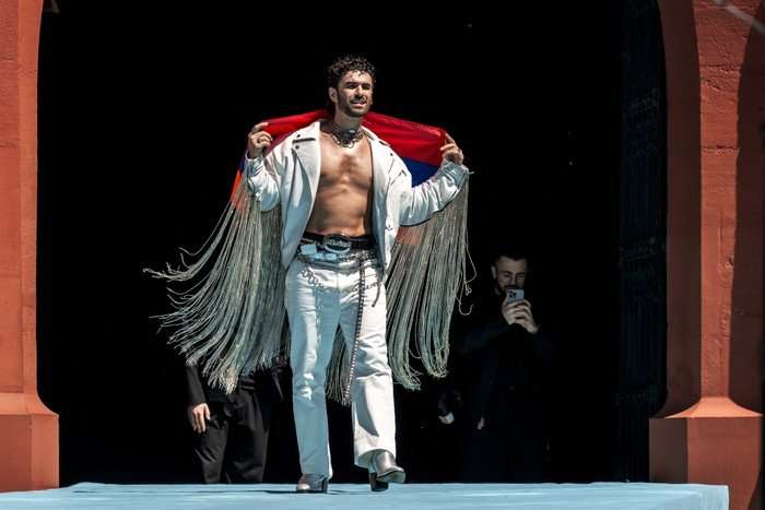 A man wearing a two piece white clothing set, holding a red cape behind him.