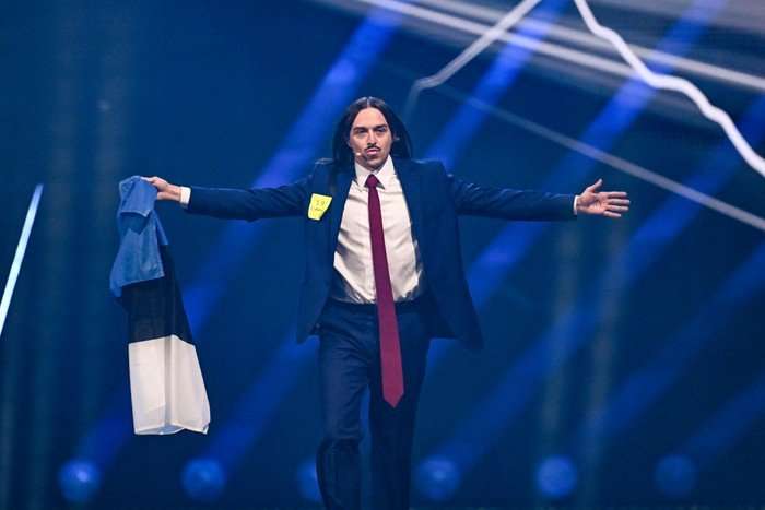 Tommy Cash walking down the stage, wearing a blue suit and long red tie. He is holding the Estonian flag with his arms stretched outwards.
