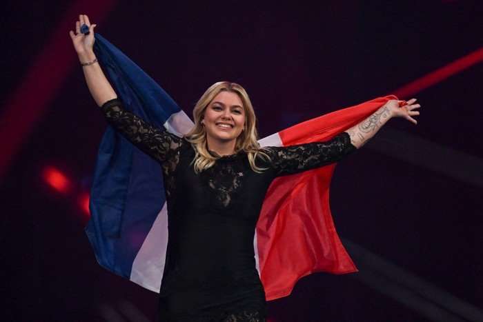 Louane wearing a black dress with her arms stretched out holding the French flag.