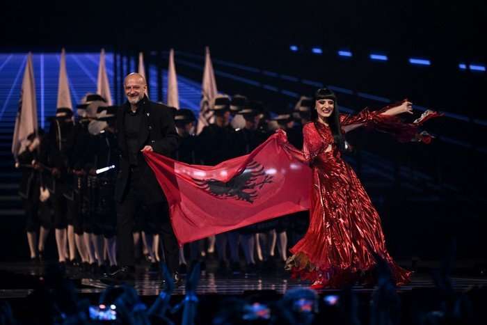 Two people stood either side of the Albanian flag, with their arms stretched out and smiling ahead.
