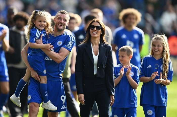 Jamie Vardy on a football pitch in Leicester City football kit, holding up a liitle girl, also in the kit, while wife Rebekah stands beside him in a black suit and sunglasses.