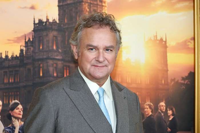 Hugh Bonneville poses in front of a picture of Downton Abbey in a grey suit with a light blue tie