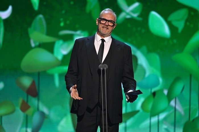 Greg Davies wearing a black suit and tie, talking into a microphone while reading out an award.
