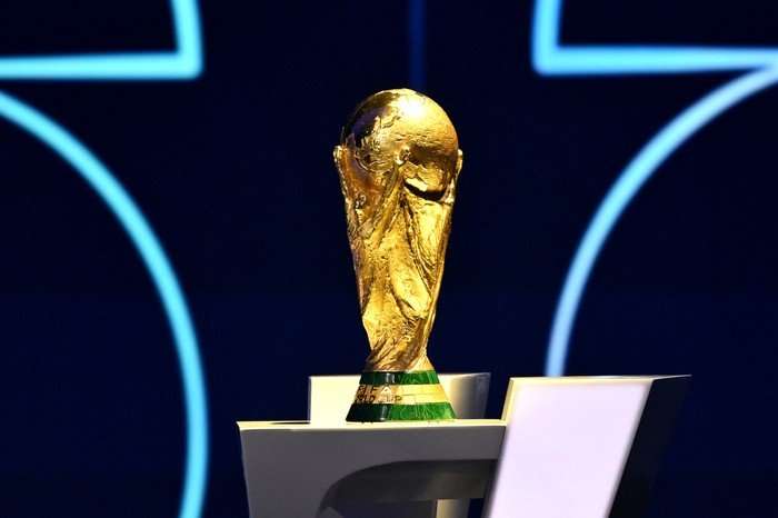 FIFA World Cup 2026 trophy Close-up photo of the FIFA World Cup trophy displayed on a white pedestal against a dark blue background with glowing geometric light patterns. The gold trophy is centered in the frame and brightly illuminated, highlighting its textured surface and green base.