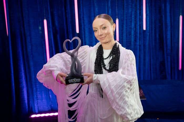 LEL&Eacute;KA holding up a trophy wearing a white dress and black beaded necklace smiling ahead.