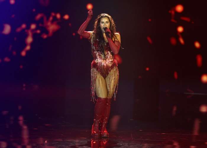 Sarah Engels singing intro a microphone on stage with arm in the air above her head looking ahead.